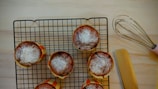 Six baked muffins with powdered sugar on top are placed on a cooling rack. A pink-handled whisk and a spatula are nearby on a light wooden surface.