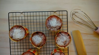Six baked muffins with powdered sugar on top are placed on a cooling rack. A pink-handled whisk and a spatula are nearby on a light wooden surface.