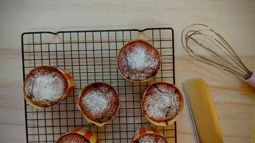 Six baked muffins with powdered sugar on top are placed on a cooling rack. A pink-handled whisk and a spatula are nearby on a light wooden surface.