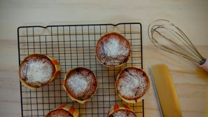 Six baked muffins with powdered sugar on top are placed on a cooling rack. A pink-handled whisk and a spatula are nearby on a light wooden surface.