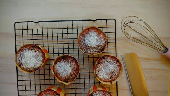 Six baked muffins with powdered sugar on top are placed on a cooling rack. A pink-handled whisk and a spatula are nearby on a light wooden surface.