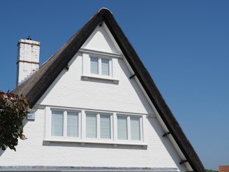 A white brick house with a steeply pitched thatched roof and a prominent chimney. The facade has several large windows with white frames, set against a bright blue sky. A small portion of leafy greenery is visible on the left side.