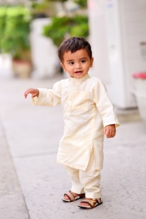 A young child with short dark hair is dressed in traditional light-colored clothing, standing on a paved pathway. The child has a cheerful expression and is wearing sandals. In the background, there is greenery with blurred foliage and a pot.