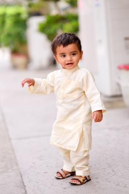 A young child with short dark hair is dressed in traditional light-colored clothing, standing on a paved pathway. The child has a cheerful expression and is wearing sandals. In the background, there is greenery with blurred foliage and a pot.