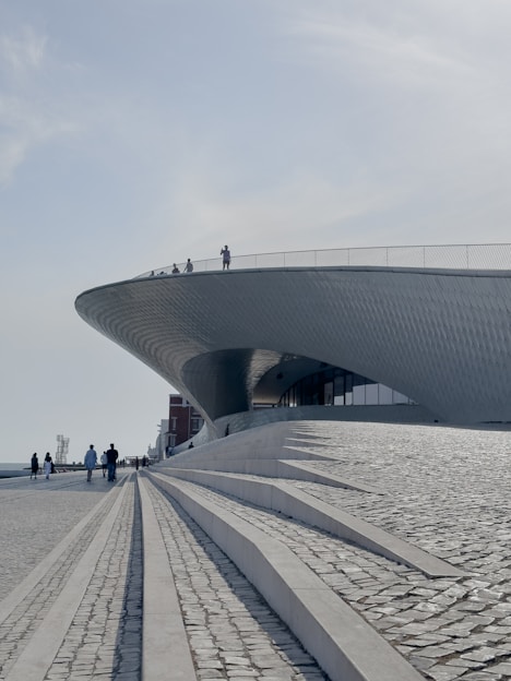 A sweeping, futuristic architectural structure with a smooth, curved roof overlooks a wide pedestrian area with cobblestone paths and steps. Several people are scattered across the scene, walking or standing, some on the roof. The sky is clear and the overall atmosphere is calm and expansive.