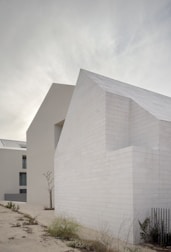 A minimalist architectural structure with clean, white block walls and a modern design is set against a cloudy sky. Sparse vegetation grows in the foreground, adding a touch of nature to the otherwise stark scene.