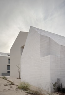 A minimalist architectural structure with clean, white block walls and a modern design is set against a cloudy sky. Sparse vegetation grows in the foreground, adding a touch of nature to the otherwise stark scene.