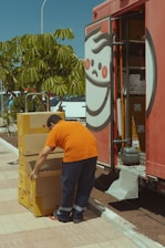 A person in an orange shirt and dark pants is stacking yellow boxes labeled with a brand logo. The person is standing near an open truck with a large logo on the side and shelving inside. Nearby, there is a green leafy tree and some parked cars.