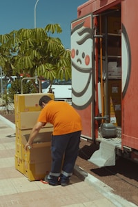 A person in an orange shirt and dark pants is stacking yellow boxes labeled with a brand logo. The person is standing near an open truck with a large logo on the side and shelving inside. Nearby, there is a green leafy tree and some parked cars.