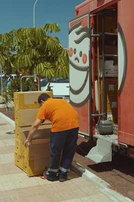 A person in an orange shirt and dark pants is stacking yellow boxes labeled with a brand logo. The person is standing near an open truck with a large logo on the side and shelving inside. Nearby, there is a green leafy tree and some parked cars.