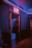 A dimly lit hallway with wooden double doors slightly ajar, revealing a room beyond. A neon sign above the doors glows with the letters 'VIP' in bright pink. The ambient lighting casts blue and pink hues onto the walls and floor, creating a moody atmosphere.