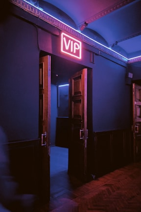 A dimly lit hallway with wooden double doors slightly ajar, revealing a room beyond. A neon sign above the doors glows with the letters 'VIP' in bright pink. The ambient lighting casts blue and pink hues onto the walls and floor, creating a moody atmosphere.