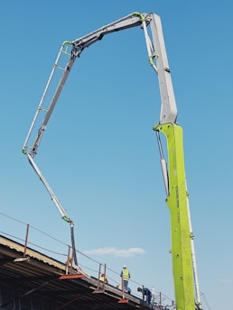 A construction site features an extended concrete pump with a bright green arm reaching high into the clear blue sky. Workers in construction gear, including hard hats and reflective vests, are positioned on a structure below, attending to various tasks. The setting suggests an active work environment on a building site.