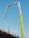 A construction site features an extended concrete pump with a bright green arm reaching high into the clear blue sky. Workers in construction gear, including hard hats and reflective vests, are positioned on a structure below, attending to various tasks. The setting suggests an active work environment on a building site.