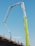 A construction site features an extended concrete pump with a bright green arm reaching high into the clear blue sky. Workers in construction gear, including hard hats and reflective vests, are positioned on a structure below, attending to various tasks. The setting suggests an active work environment on a building site.