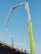 A construction site features an extended concrete pump with a bright green arm reaching high into the clear blue sky. Workers in construction gear, including hard hats and reflective vests, are positioned on a structure below, attending to various tasks. The setting suggests an active work environment on a building site.