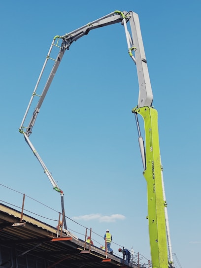 A construction site features an extended concrete pump with a bright green arm reaching high into the clear blue sky. Workers in construction gear, including hard hats and reflective vests, are positioned on a structure below, attending to various tasks. The setting suggests an active work environment on a building site.