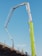 A construction site features an extended concrete pump with a bright green arm reaching high into the clear blue sky. Workers in construction gear, including hard hats and reflective vests, are positioned on a structure below, attending to various tasks. The setting suggests an active work environment on a building site.