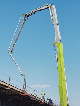 A construction site features an extended concrete pump with a bright green arm reaching high into the clear blue sky. Workers in construction gear, including hard hats and reflective vests, are positioned on a structure below, attending to various tasks. The setting suggests an active work environment on a building site.