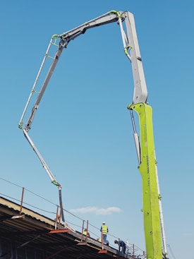 A construction site features an extended concrete pump with a bright green arm reaching high into the clear blue sky. Workers in construction gear, including hard hats and reflective vests, are positioned on a structure below, attending to various tasks. The setting suggests an active work environment on a building site.