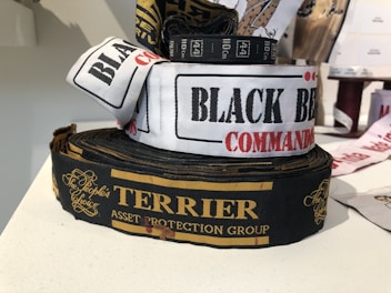 Several rolls of woven label tapes are displayed on a surface. The labels include inscriptions in various fonts and colors, such as 'BLACK BEAR COMMANDO' on a white background and 'TERRIER ASSET PROTECTION GROUP' on a black background with gold lettering. Other labels are partially visible in the background.