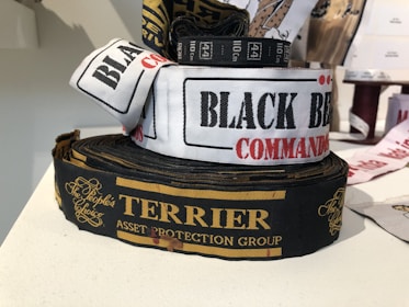 Several rolls of woven label tapes are displayed on a surface. The labels include inscriptions in various fonts and colors, such as 'BLACK BEAR COMMANDO' on a white background and 'TERRIER ASSET PROTECTION GROUP' on a black background with gold lettering. Other labels are partially visible in the background.
