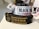 Several rolls of woven label tapes are displayed on a surface. The labels include inscriptions in various fonts and colors, such as 'BLACK BEAR COMMANDO' on a white background and 'TERRIER ASSET PROTECTION GROUP' on a black background with gold lettering. Other labels are partially visible in the background.
