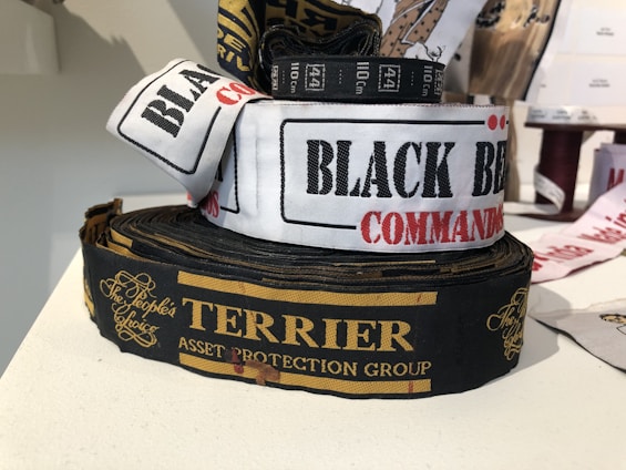 Several rolls of woven label tapes are displayed on a surface. The labels include inscriptions in various fonts and colors, such as 'BLACK BEAR COMMANDO' on a white background and 'TERRIER ASSET PROTECTION GROUP' on a black background with gold lettering. Other labels are partially visible in the background.