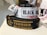 Several rolls of woven label tapes are displayed on a surface. The labels include inscriptions in various fonts and colors, such as 'BLACK BEAR COMMANDO' on a white background and 'TERRIER ASSET PROTECTION GROUP' on a black background with gold lettering. Other labels are partially visible in the background.