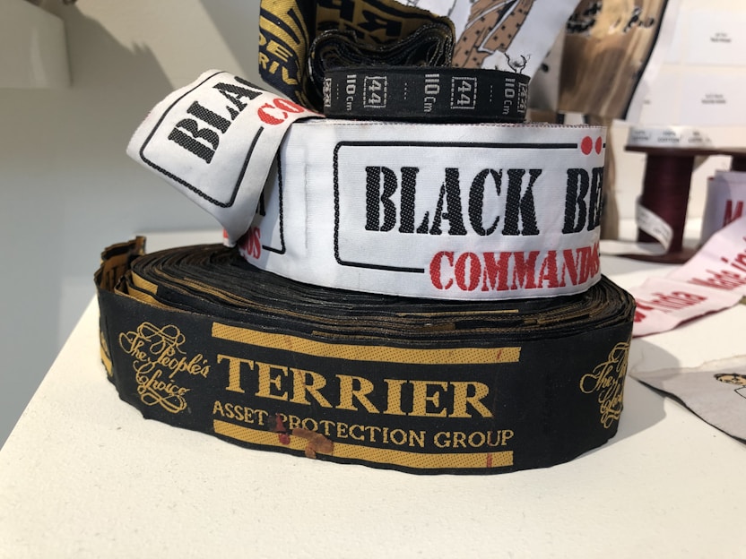 Several rolls of woven label tapes are displayed on a surface. The labels include inscriptions in various fonts and colors, such as 'BLACK BEAR COMMANDO' on a white background and 'TERRIER ASSET PROTECTION GROUP' on a black background with gold lettering. Other labels are partially visible in the background.