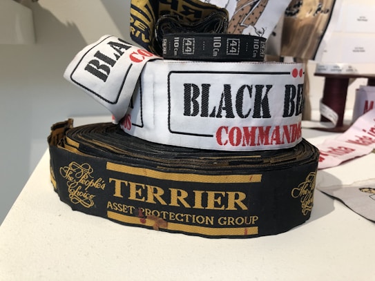 Several rolls of woven label tapes are displayed on a surface. The labels include inscriptions in various fonts and colors, such as 'BLACK BEAR COMMANDO' on a white background and 'TERRIER ASSET PROTECTION GROUP' on a black background with gold lettering. Other labels are partially visible in the background.