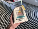 A hand holds a small beige drawstring bag featuring a colorful diagram resembling Maslow's Pyramid of Needs. The pyramid has sections in yellow, pink, and teal. The background consists of a checkered geometric-patterned floor with a table on the left side.