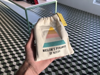 A hand holds a small beige drawstring bag featuring a colorful diagram resembling Maslow's Pyramid of Needs. The pyramid has sections in yellow, pink, and teal. The background consists of a checkered geometric-patterned floor with a table on the left side.