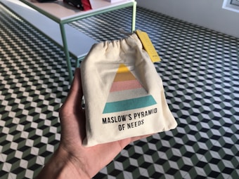 A hand holds a small beige drawstring bag featuring a colorful diagram resembling Maslow's Pyramid of Needs. The pyramid has sections in yellow, pink, and teal. The background consists of a checkered geometric-patterned floor with a table on the left side.