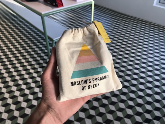 A hand holds a small beige drawstring bag featuring a colorful diagram resembling Maslow's Pyramid of Needs. The pyramid has sections in yellow, pink, and teal. The background consists of a checkered geometric-patterned floor with a table on the left side.
