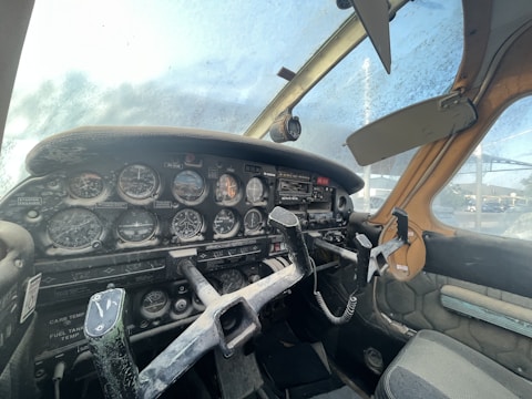 A vintage aircraft cockpit with a variety of analog dials and controls. The panel is filled with gauges, switches, and a yoke. The windshield appears foggy or dirty, indicating the cockpit might not have been used recently. Sun visors are attached above the windshield, and the seats have a worn fabric appearance.