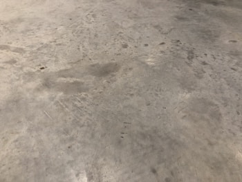 A concrete floor with various stains and scuff marks, showing a worn and used appearance. The surface has multiple shades of gray with darker spots scattered throughout.