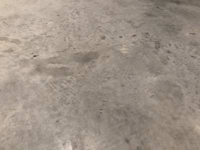 A concrete floor with various stains and scuff marks, showing a worn and used appearance. The surface has multiple shades of gray with darker spots scattered throughout.