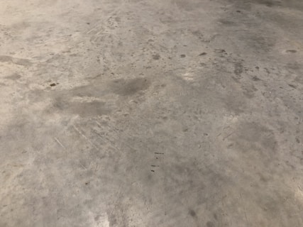 A concrete floor with various stains and scuff marks, showing a worn and used appearance. The surface has multiple shades of gray with darker spots scattered throughout.