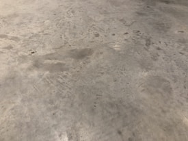A concrete floor with various stains and scuff marks, showing a worn and used appearance. The surface has multiple shades of gray with darker spots scattered throughout.