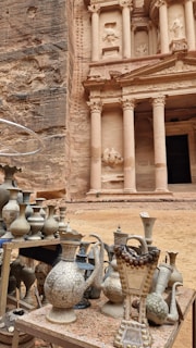 An ancient carved stone facade with tall columns stands majestically in the background. In the foreground, intricately designed metal vases and jugs are displayed on a table, suggesting a market or display of traditional crafts. The stonework and objects evoke a sense of history and cultural richness.