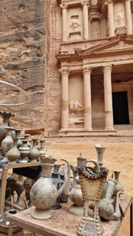 An ancient carved stone facade with tall columns stands majestically in the background. In the foreground, intricately designed metal vases and jugs are displayed on a table, suggesting a market or display of traditional crafts. The stonework and objects evoke a sense of history and cultural richness.
