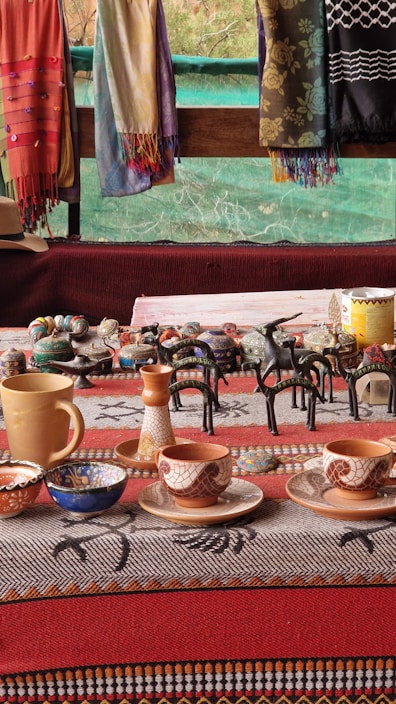 A collection of handcrafted ceramic cups, bowls, and pottery pieces displayed on a table with a patterned cloth. Colorful scarves hang in the background, adding vibrancy to the scene. The pottery includes various intricate designs, with a focus on earthy tones and patterns.