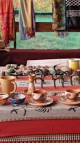 A collection of handcrafted ceramic cups, bowls, and pottery pieces displayed on a table with a patterned cloth. Colorful scarves hang in the background, adding vibrancy to the scene. The pottery includes various intricate designs, with a focus on earthy tones and patterns.