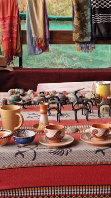 A collection of handcrafted ceramic cups, bowls, and pottery pieces displayed on a table with a patterned cloth. Colorful scarves hang in the background, adding vibrancy to the scene. The pottery includes various intricate designs, with a focus on earthy tones and patterns.