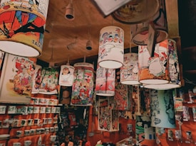 A ceiling display features hanging lampshades adorned with colorful and whimsical cartoon illustrations. The shelves below are stocked with a variety of mugs and other merchandise featuring similar cartoon art. The overall setting seems to be a gift shop or a similar retail space filled with vibrant and humorous cartoon-themed products.