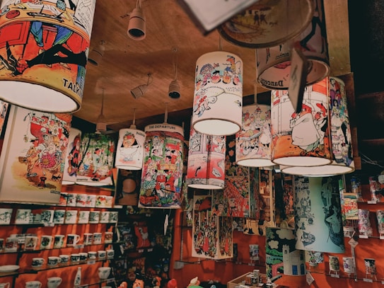A ceiling display features hanging lampshades adorned with colorful and whimsical cartoon illustrations. The shelves below are stocked with a variety of mugs and other merchandise featuring similar cartoon art. The overall setting seems to be a gift shop or a similar retail space filled with vibrant and humorous cartoon-themed products.
