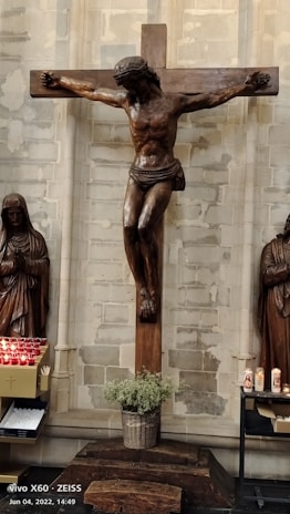 A wooden crucifix with a depiction of Jesus hangs prominently against a stone wall. On either side, there are statues in robes, likely religious figures. A basket with greenery is placed beneath the crucifix. To the left, an array of red prayer candles and a box of votive candles are visible. On the right, more candles are lit, including one with an image on the front.