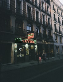A dimly lit street scene in front of a café bar with a neon sign reading 'La Vera,' situated at the bottom of a building with classical architecture. People are walking on the sidewalk along the storefront.