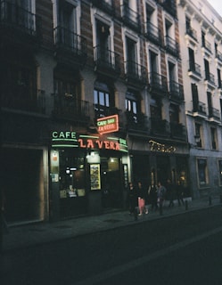 A dimly lit street scene in front of a café bar with a neon sign reading 'La Vera,' situated at the bottom of a building with classical architecture. People are walking on the sidewalk along the storefront.